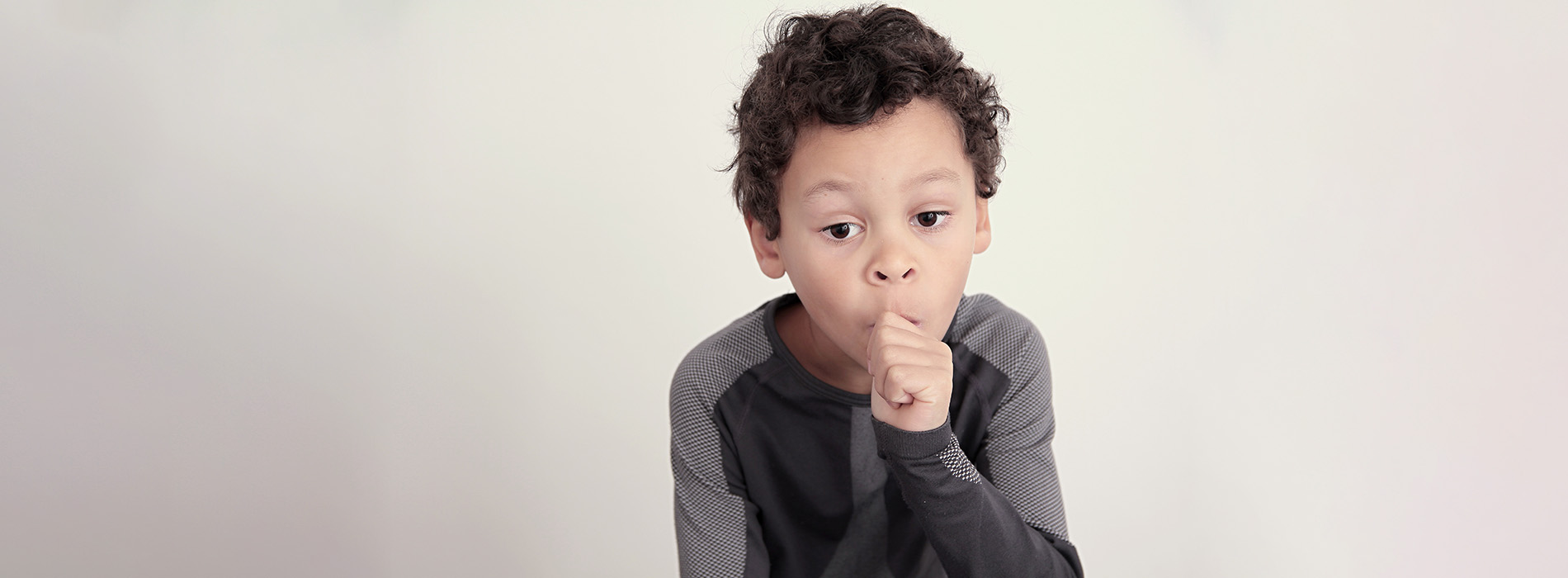 The image shows a young boy with curly hair wearing a dark top, making a playful gesture with his hand near his mouth while standing against a plain background.