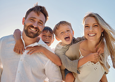 The image shows a family of four with two adults and two children, smiling and posing together outdoors during daytime.