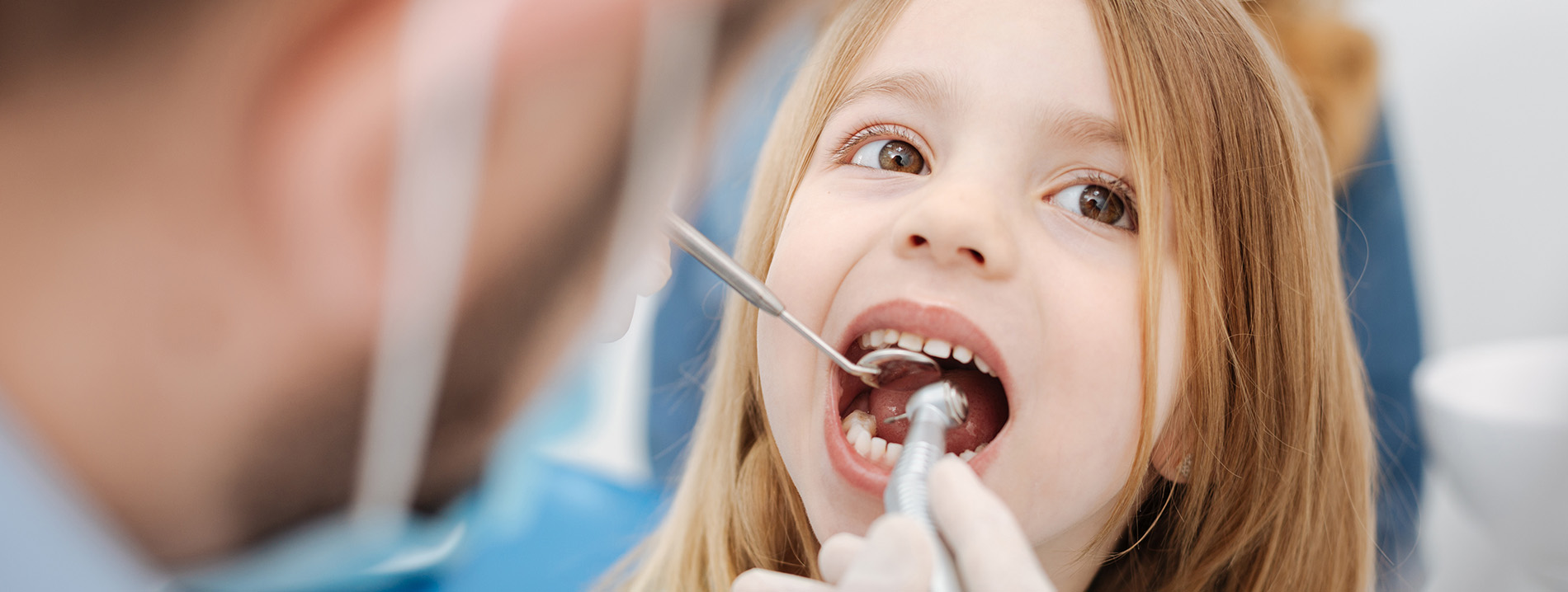 A young girl with her mouth open wide, being attended to by a dental professional who is holding a mirror while seated at a chair.