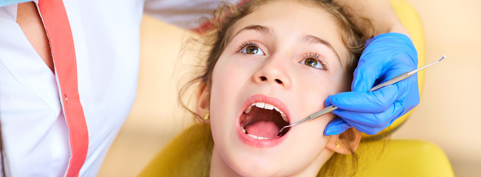 A young girl receiving dental treatment with a dentist s hand visible, wearing a blue glove and holding a dental instrument.