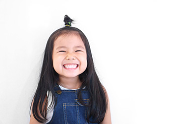 The image shows a young child with a joyful expression, smiling at the camera, wearing denim overalls and a hair accessory.