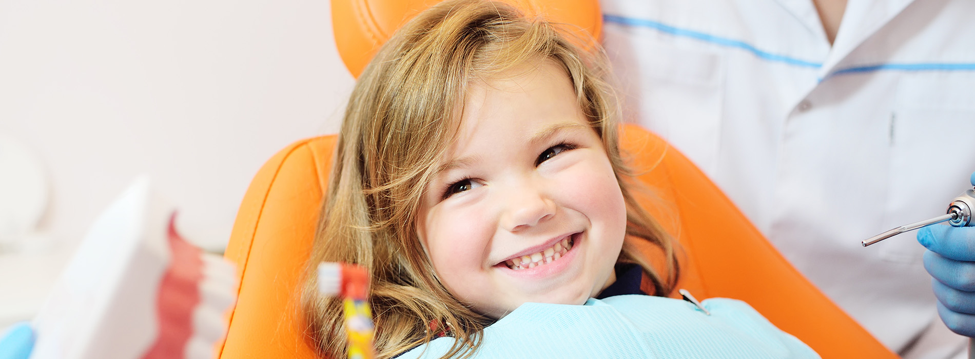 The image depicts a young child sitting in an orange chair with a smile on their face, surrounded by medical professionals wearing white coats and blue aprons, indicating a hospital setting.