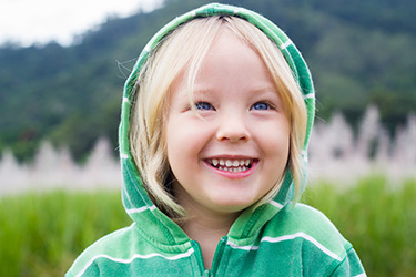 A young child with blonde hair wearing a green hoodie, smiling at the camera while standing outdoors during daylight.