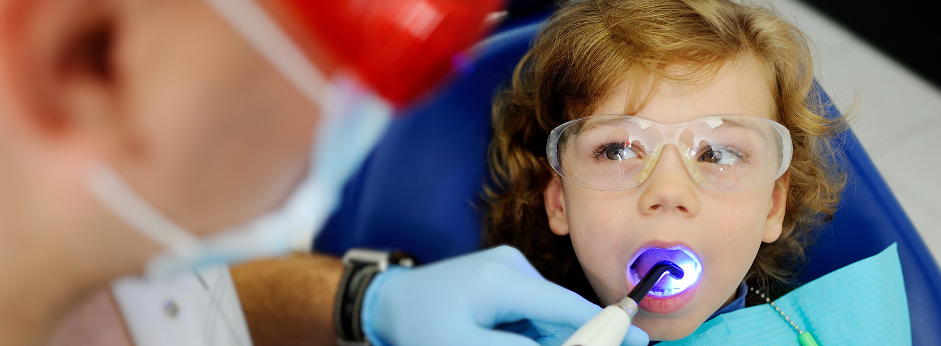 The image shows a young child wearing safety goggles and a dental mouthguard, with an adult in the background who appears to be a dentist or dental hygienist, examining the child s teeth using a dental mirror.