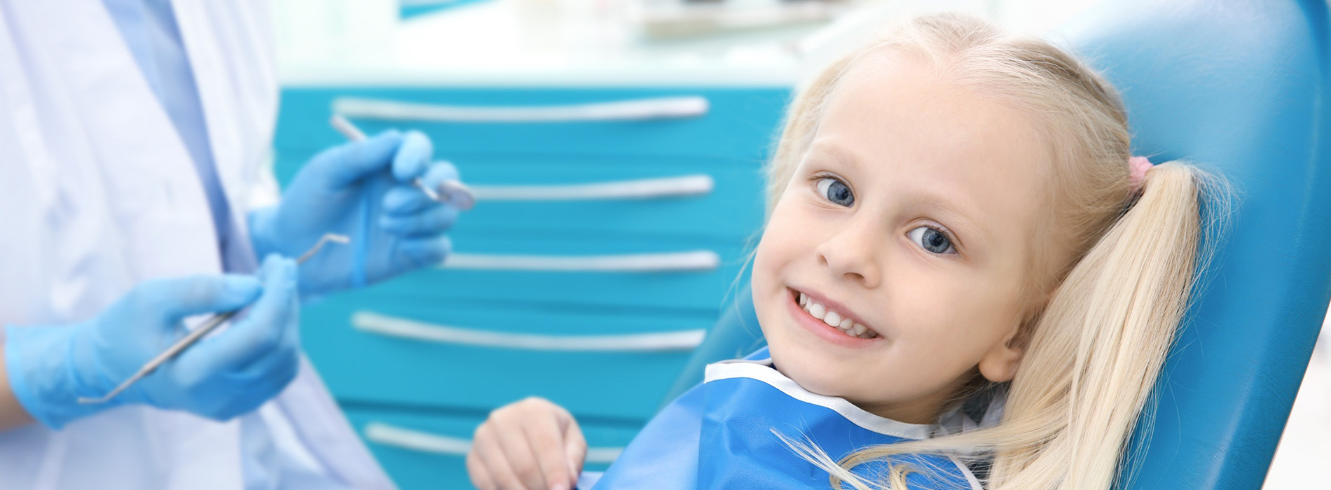 The image shows a young child sitting in a dentist s chair with a smiling expression, receiving dental care from a professional wearing gloves and a blue uniform, with other dental equipment visible in the background.