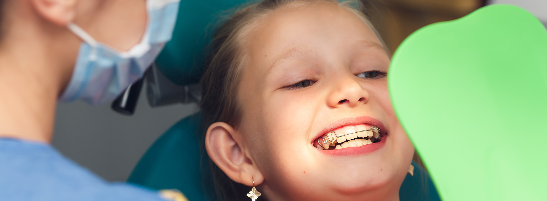 The image shows a young girl with red lipstick sitting in a dental chair, smiling at the camera, while a dentist is performing a procedure on her teeth.