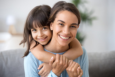 A woman hugging a young girl, both smiling, with a warm embrace against a domestic backdrop.