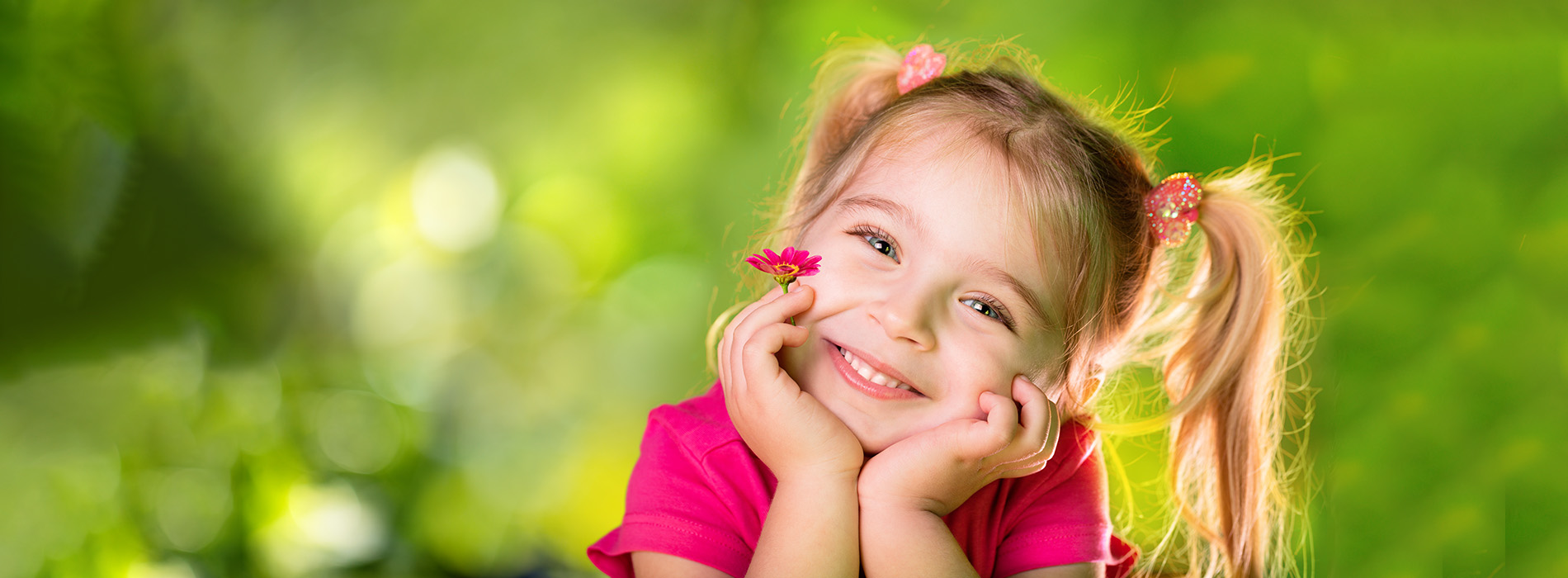 The image shows a young girl with her hand on her chin, smiling at the camera, set against a blurred background of greenery.