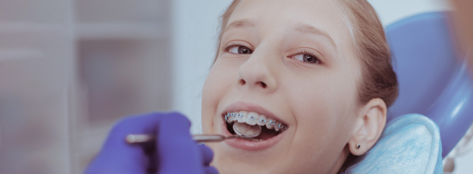 The image shows a young girl with braces undergoing dental treatment, sitting in a dental chair while smiling at the camera.