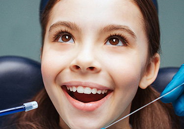 The image features a young girl with braces receiving dental care, smiling broadly while holding her mouth open for the dentist s examination.