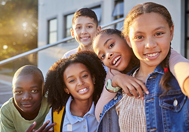 The image shows a group of children posing together outdoors during daylight, with a school building in the background.
