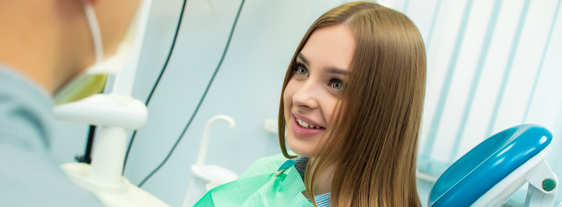 The image shows a woman with long hair sitting in front of a dental chair, smiling at the camera, while a dentist stands behind her.