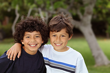 The image shows two young boys posing together with smiles, standing outdoors under a tree, with a blurred background indicating a park setting.