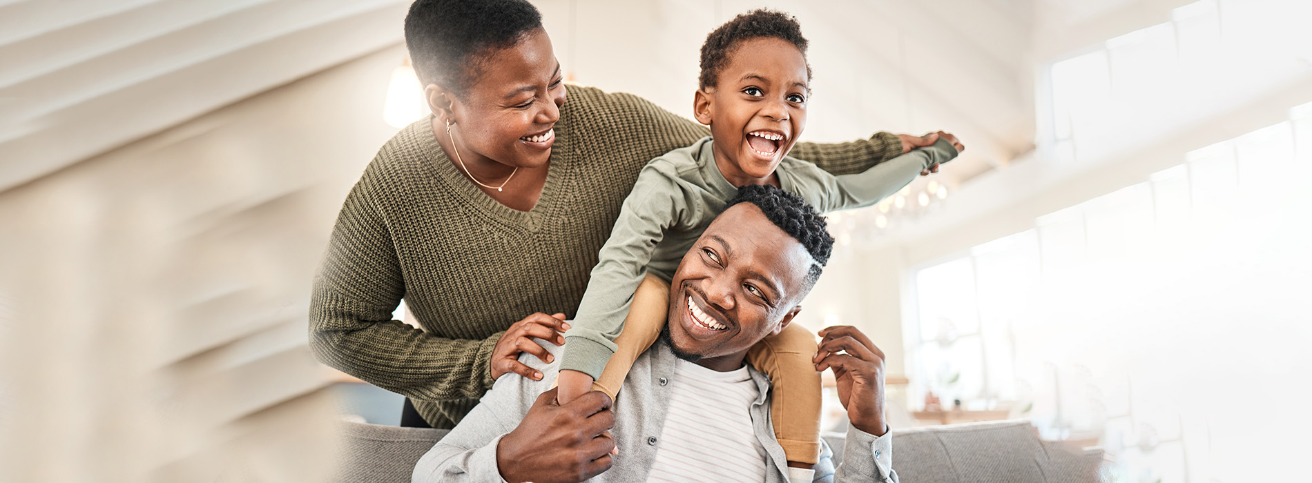 The image depicts a family of four, with two adults and two children, smiling and posing together for a photograph. They appear to be indoors, possibly in a home setting, with one adult holding a child on their hip.