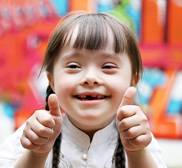 The image shows a young child with a joyful expression, thumbs up gesture, and big smile, wearing a white shirt and standing outdoors against a colorful background.