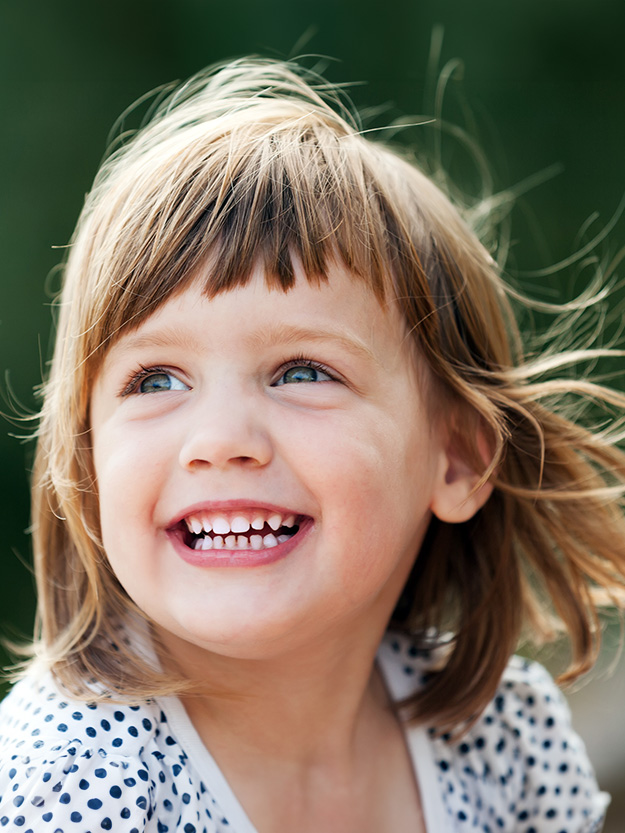 A young girl with blonde hair and blue eyes smiles at the camera outdoors.