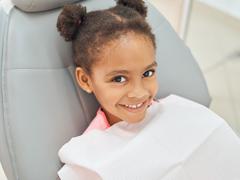 A young girl with dark hair is sitting in a dental chair, smiling at the camera.