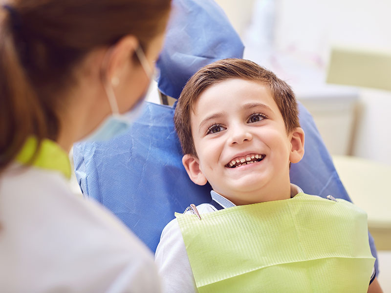 The image shows a young child sitting in a dental chair with a smile, receiving dental care from an adult wearing a blue surgical gown and a white face mask.