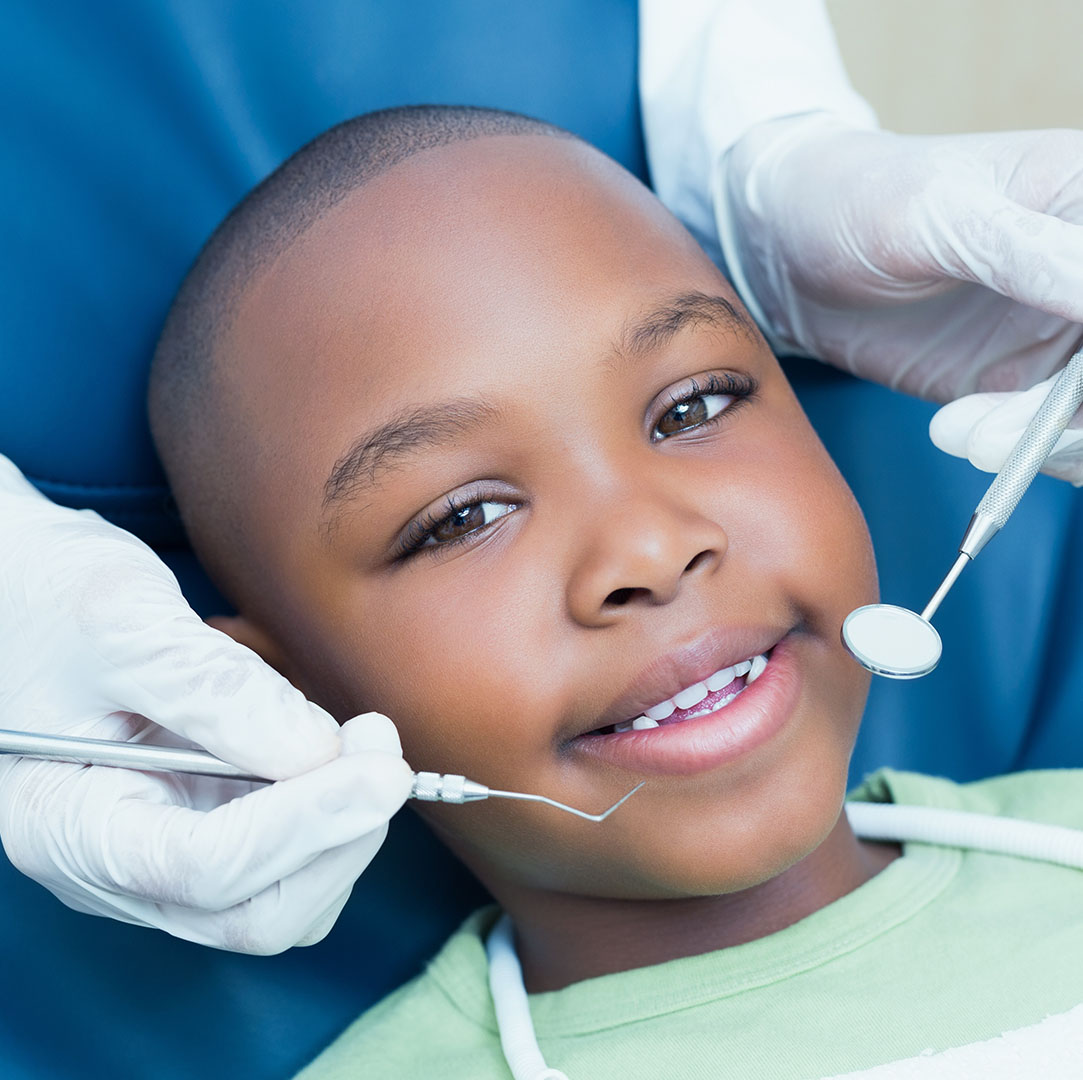 The image shows a young boy receiving dental care  he is seated in a dental chair with a smiling expression, wearing a green shirt, and being attended by a dentist who appears to be adjusting his braces or preparing him for a procedure.