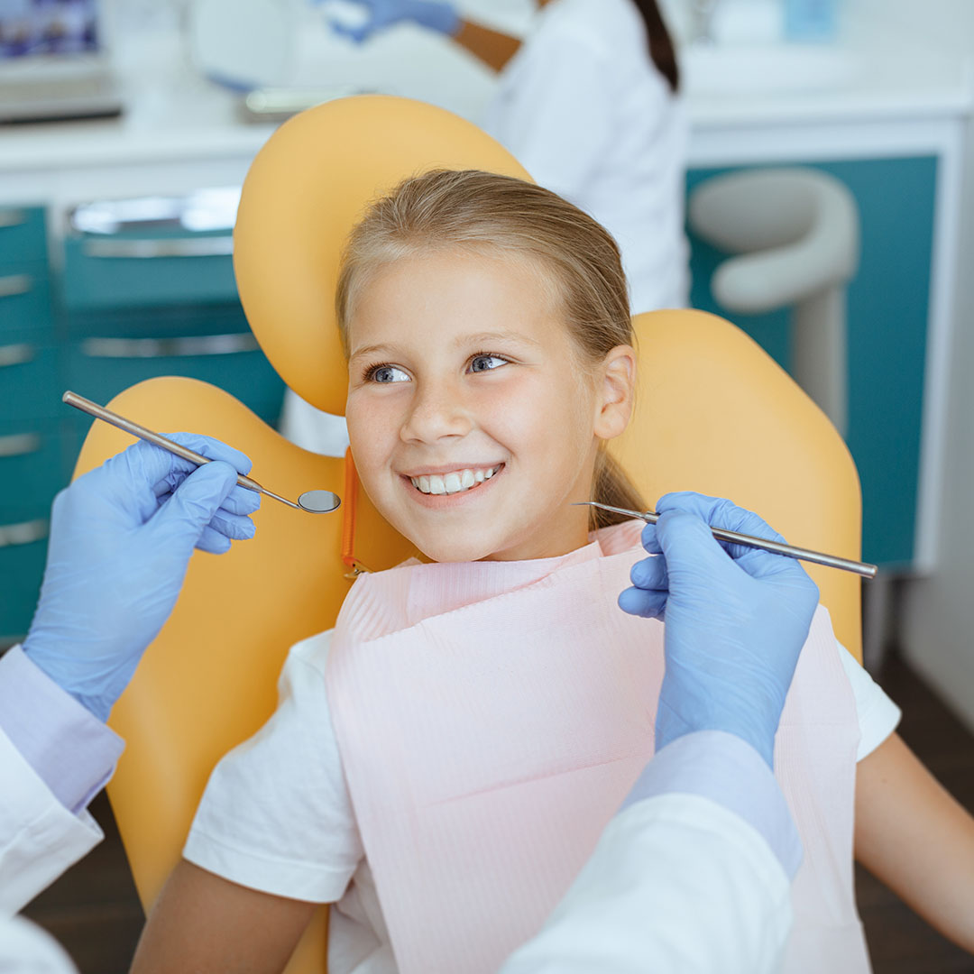 The image shows a young girl receiving dental care with a smile on her face, surrounded by dental professionals wearing blue gloves and white coats, in an office setting with dental equipment visible.